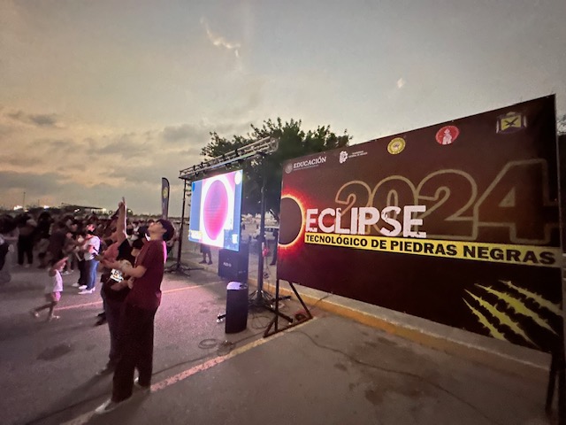 People gathered outside around a giant screen and a billboard reading “2024 Eclipse: Tecnologico de Piedras Negras”