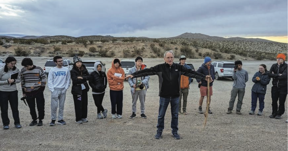 Photo of part of a large circle of students in a mostly deserted parking lot. Prof. Kirschvink stands in the center, spreading his arms wide, cane in one hand