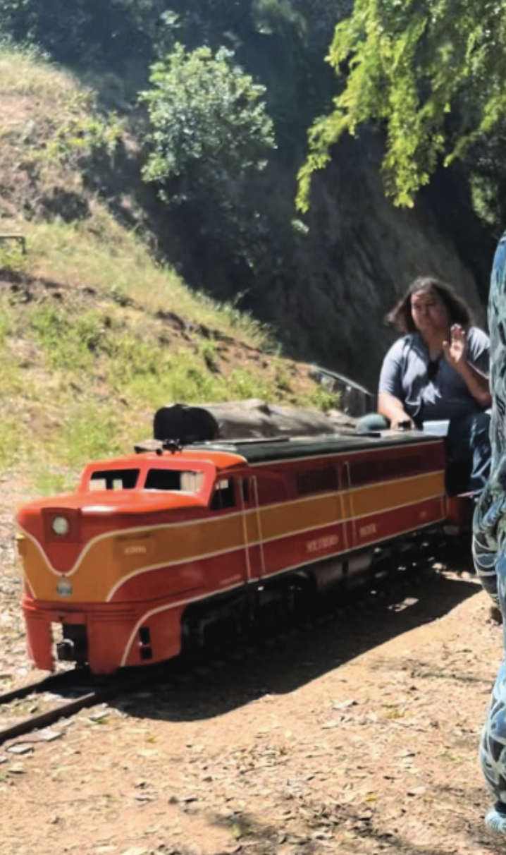 Photo of a miniature train running on a track embedded in the ground. One person is sitting in front and waving at the camera.
